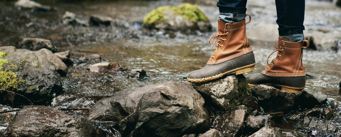 A person wearing brown waterproof boots and blue jeans walking on rocks across a river