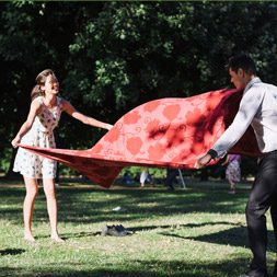 A couple unfolds a red blanket on the grass in a park.